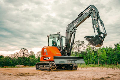 Orange Kubota mini excavator with extended boom on sandy construction site surrounded by trees
