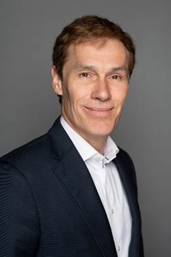 Professional headshot of a man wearing a dark blazer and light blue striped shirt against a gray background