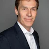 Professional headshot of a man wearing a dark blazer and light blue striped shirt against a gray background