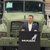 Man in business attire speaking at Mack Trucks podium in front of military truck