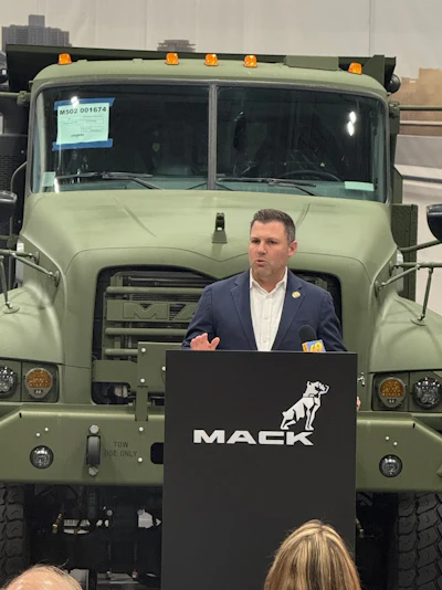 Man in business attire speaking at Mack Trucks podium in front of military truck