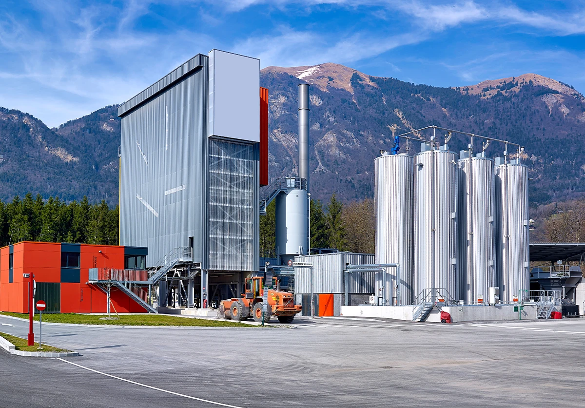 Industrial manufacturing facility with red building, silver storage tanks, and orange equipment against mountain backdrop