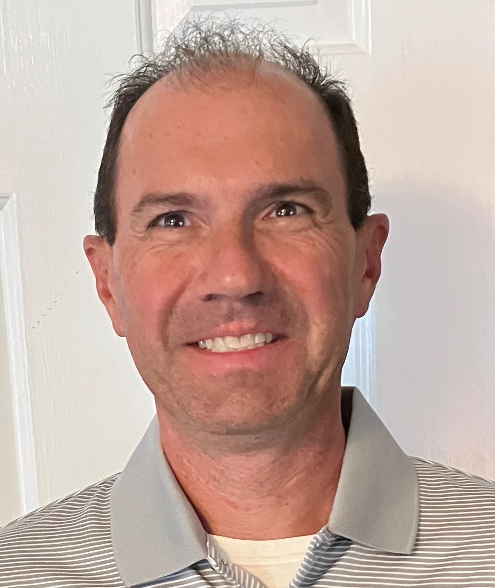 Smiling man with receding dark hair wearing a striped gray and white collared shirt against a white interior background