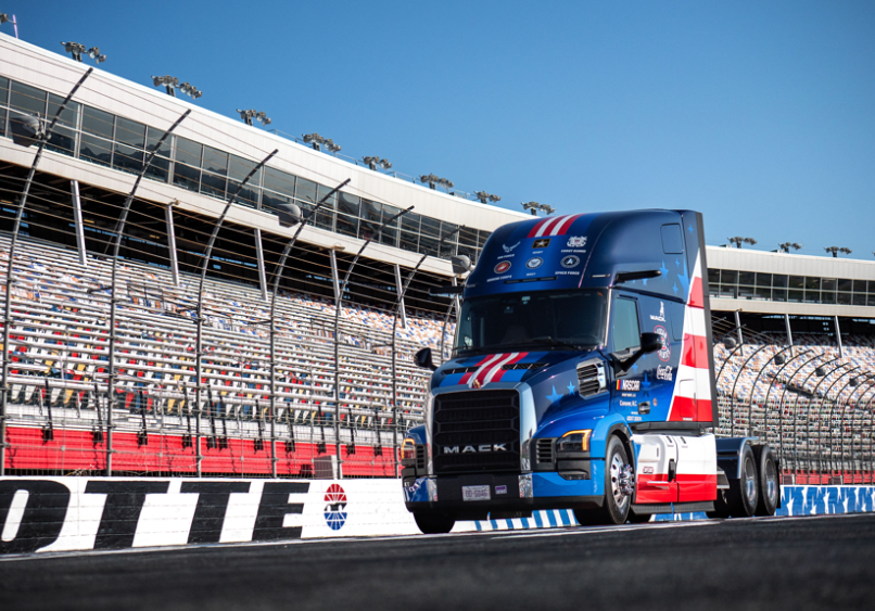 Blue and red Mack truck on racing track with stadium grandstands in background