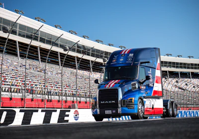 Blue and red Mack truck on racing track with stadium grandstands in background