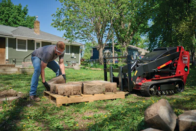 Man loading sod rolls onto a red Toro compact track loader in a residential yard with trees and houses.