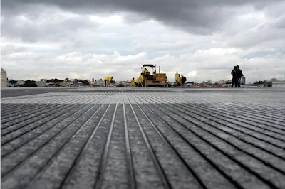 Construction crew operating heavy machinery on a large flat surface with parallel ridged patterns under cloudy sky.