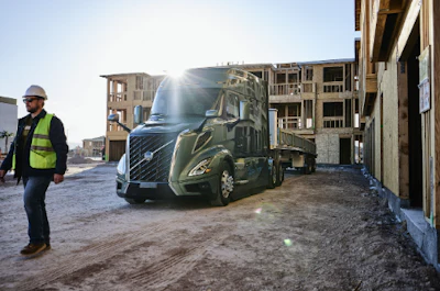 Construction worker in safety gear observing a blue Volvo semi-truck at an active building site