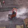 Red vibratory roller compactor creating dust clouds while compacting asphalt on construction site, aerial view