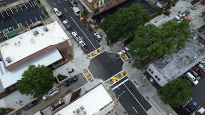 Aerial view of urban street intersection with parked cars, white-roofed buildings, trees, and yellow parking space markings