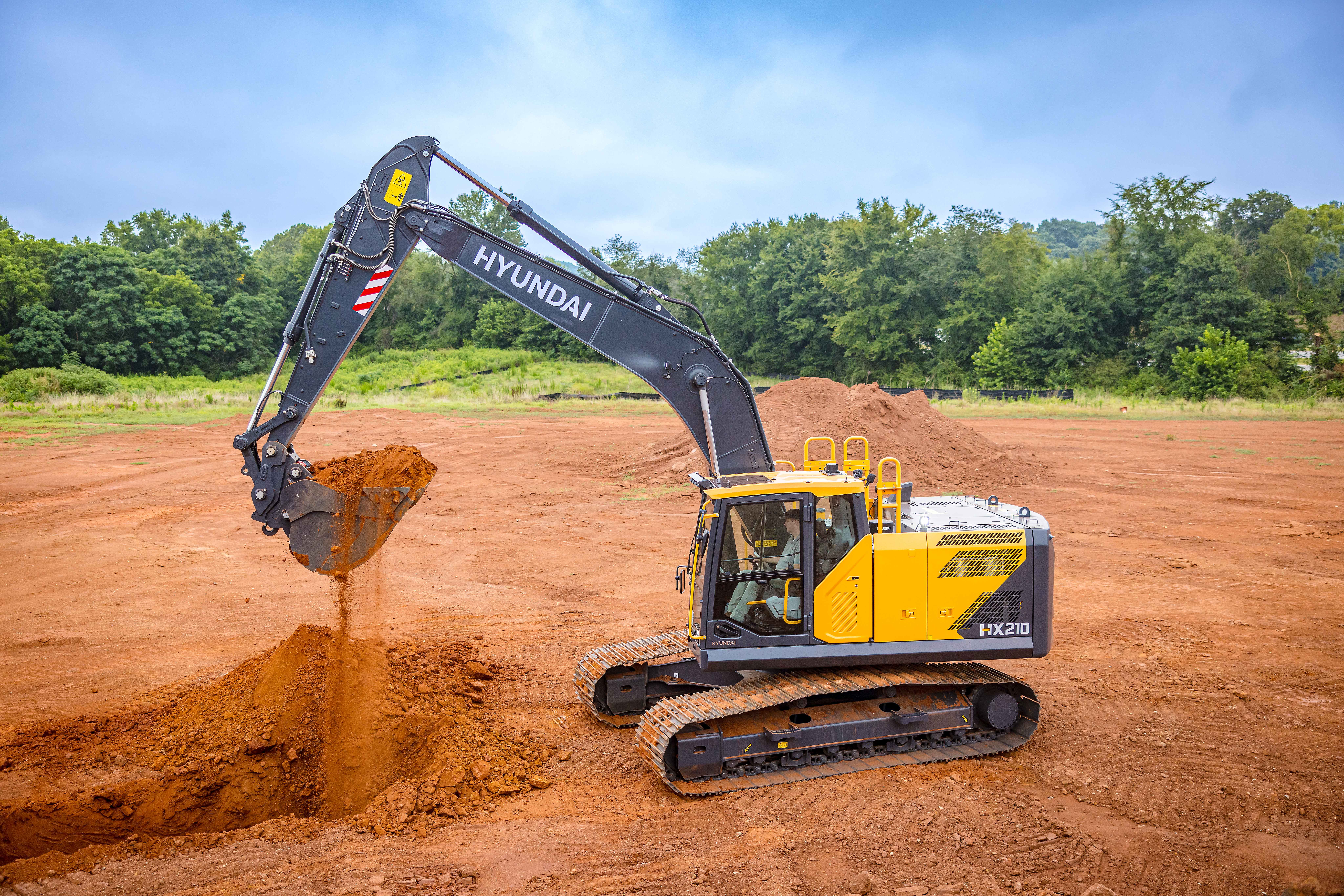 Yellow and black Hyundai excavator digging red soil at a construction site with trees in background