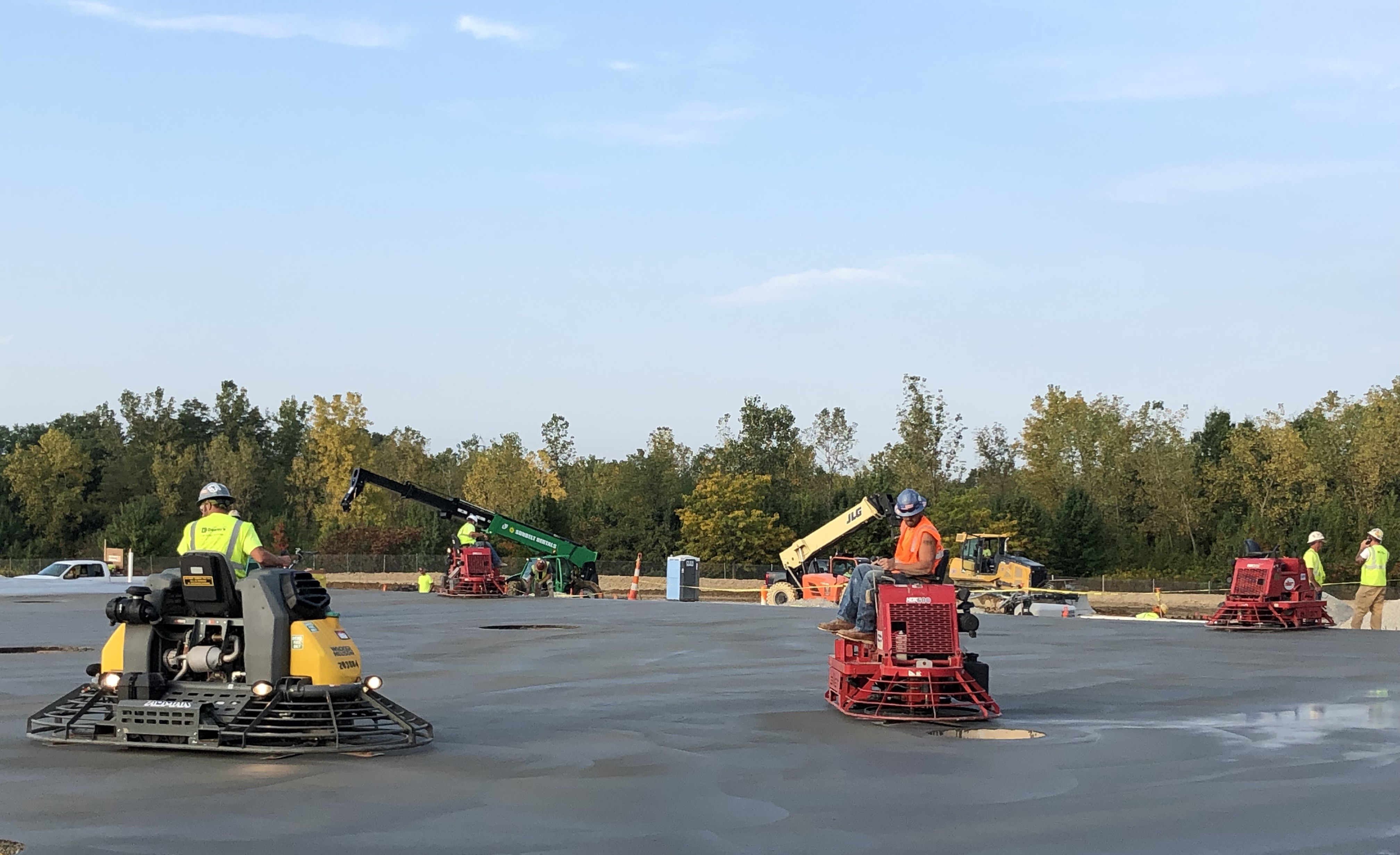 Construction workers operating concrete finishing equipment on a large paved surface with machinery and safety equipment visible in background.