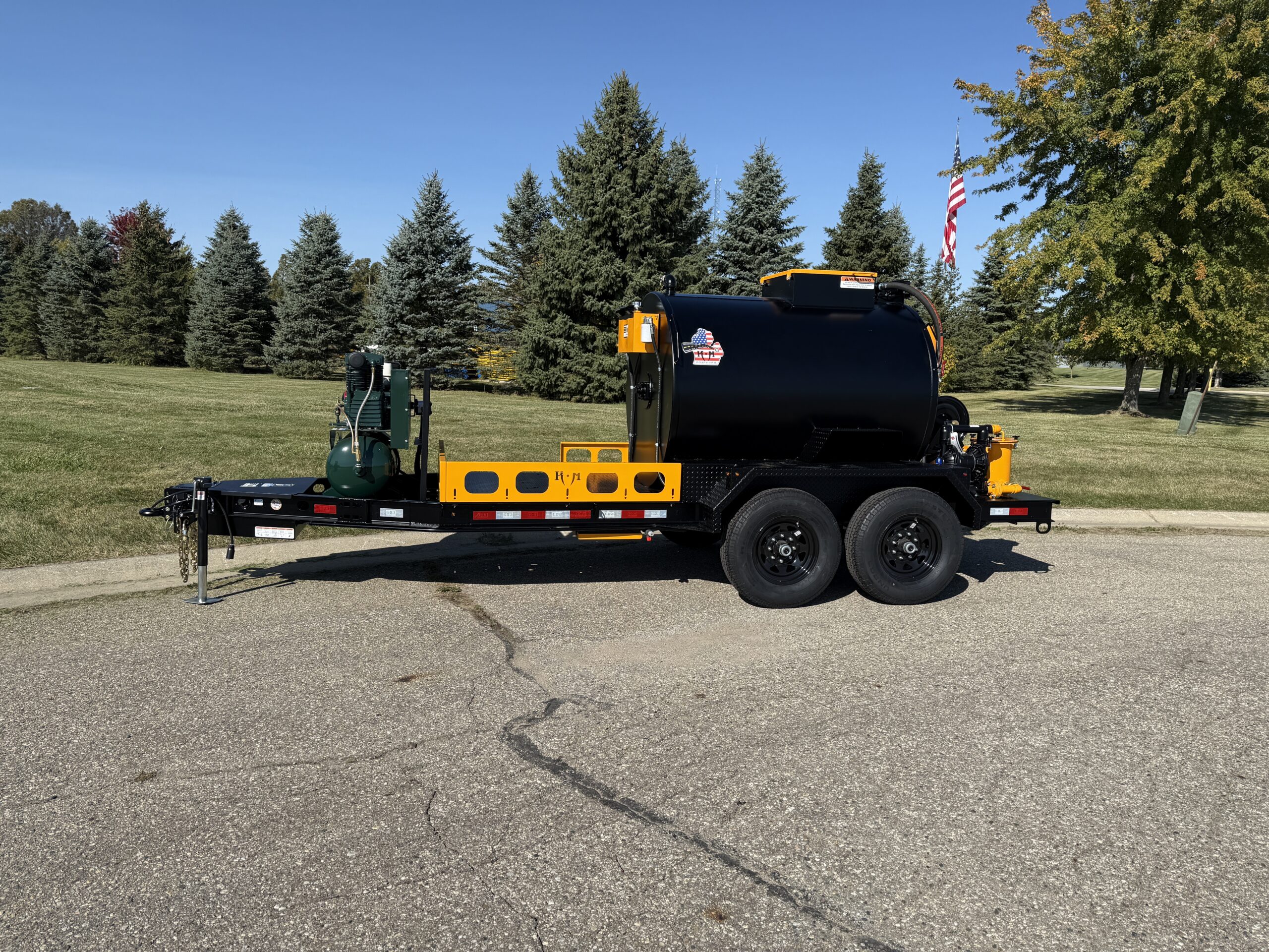 Yellow and black dual-axle asphalt tanker truck parked on pavement with evergreen trees and American flag in background