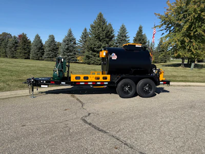 Yellow and black dual-axle asphalt tanker truck parked on pavement with evergreen trees and American flag in background