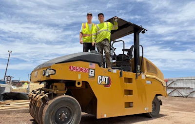 Two construction workers in yellow safety vests stand on a yellow Caterpillar JG5C65 compactor at a job site