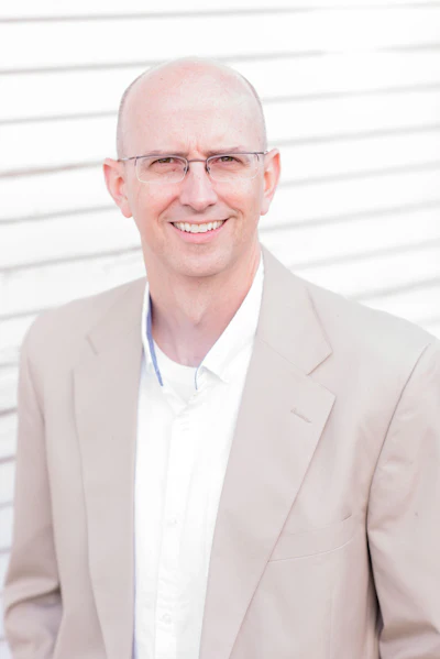 Professional headshot of a smiling man wearing glasses and beige blazer against white background