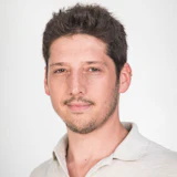 Professional headshot of a man with dark hair and facial hair wearing a light-colored collared shirt against a neutral background