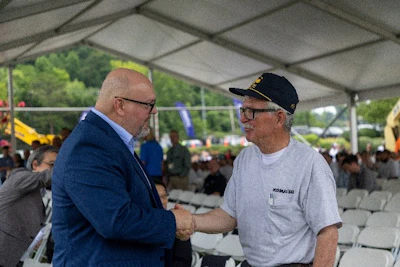 Two men shake hands at an outdoor event under a tent with spectators in background