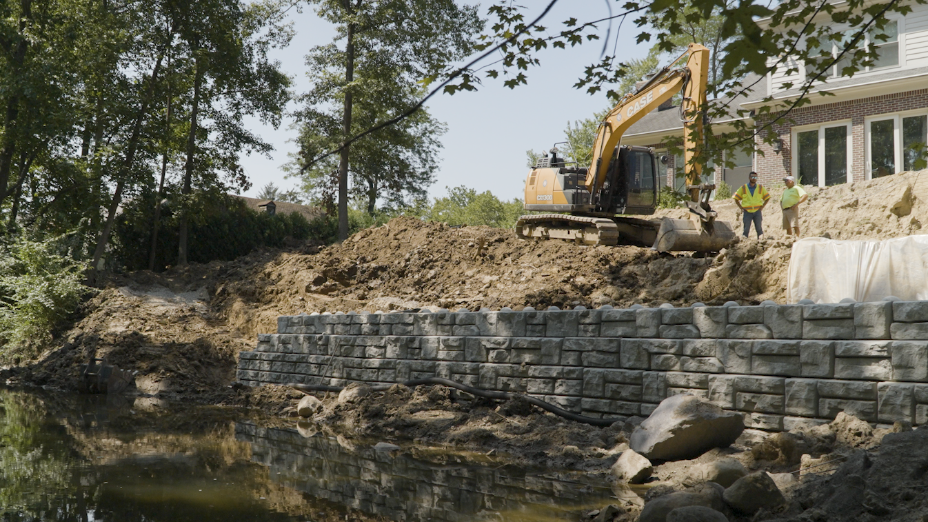 Construction site with yellow excavator building retaining wall beside residential home