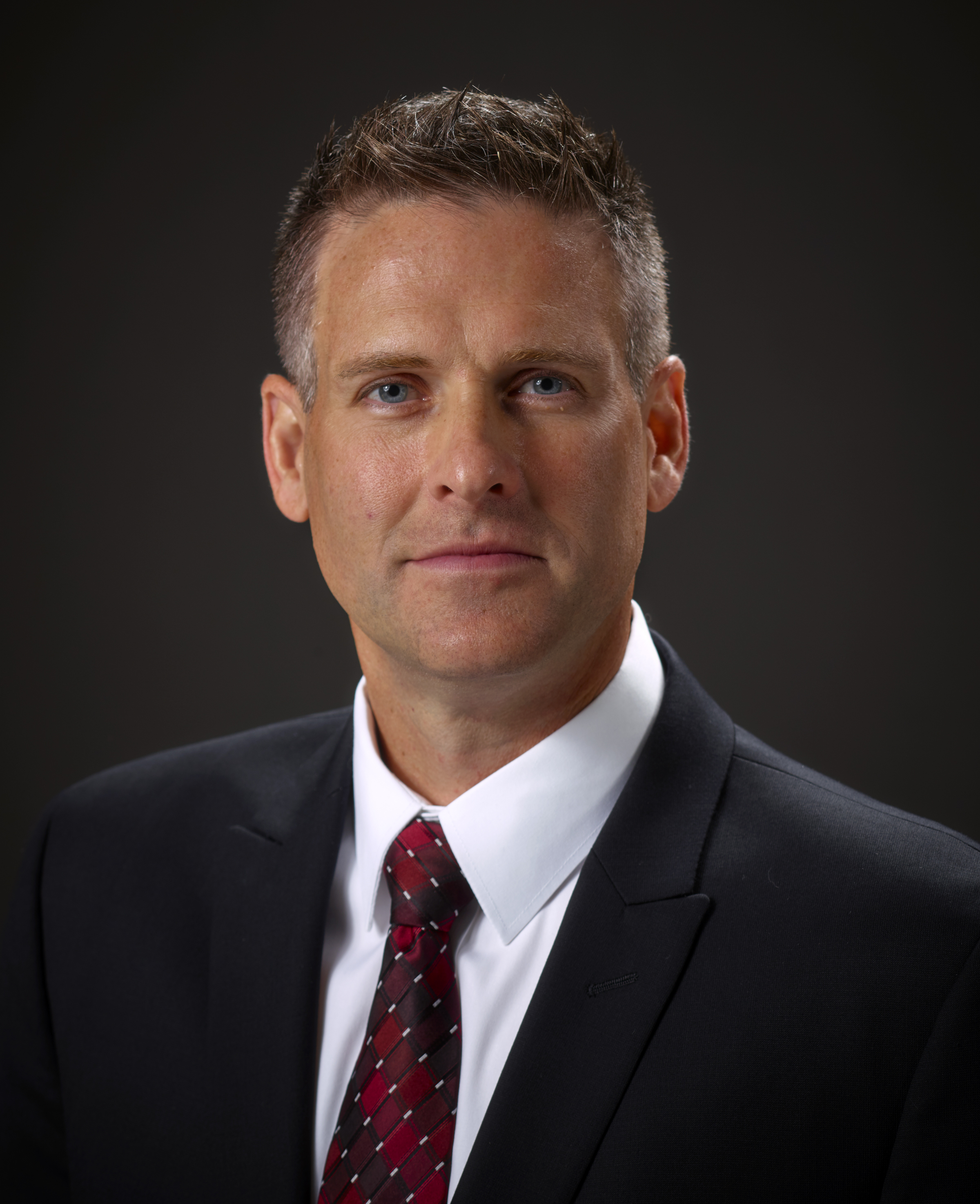 Professional headshot of a man in dark suit and burgundy tie against dark background