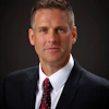 Professional headshot of a man in dark suit and burgundy tie against dark background