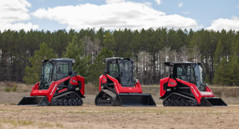 Three red and black tracked skid steer loaders lined up in a field with forest background