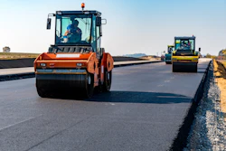 Road construction crew operating orange and yellow asphalt rollers on a newly paved highway under clear skies.