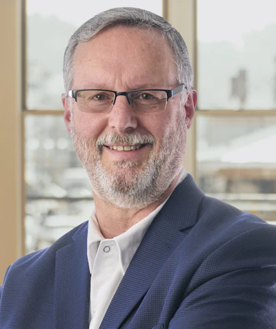 Professional headshot of a bearded man wearing glasses and navy blazer in modern office setting
