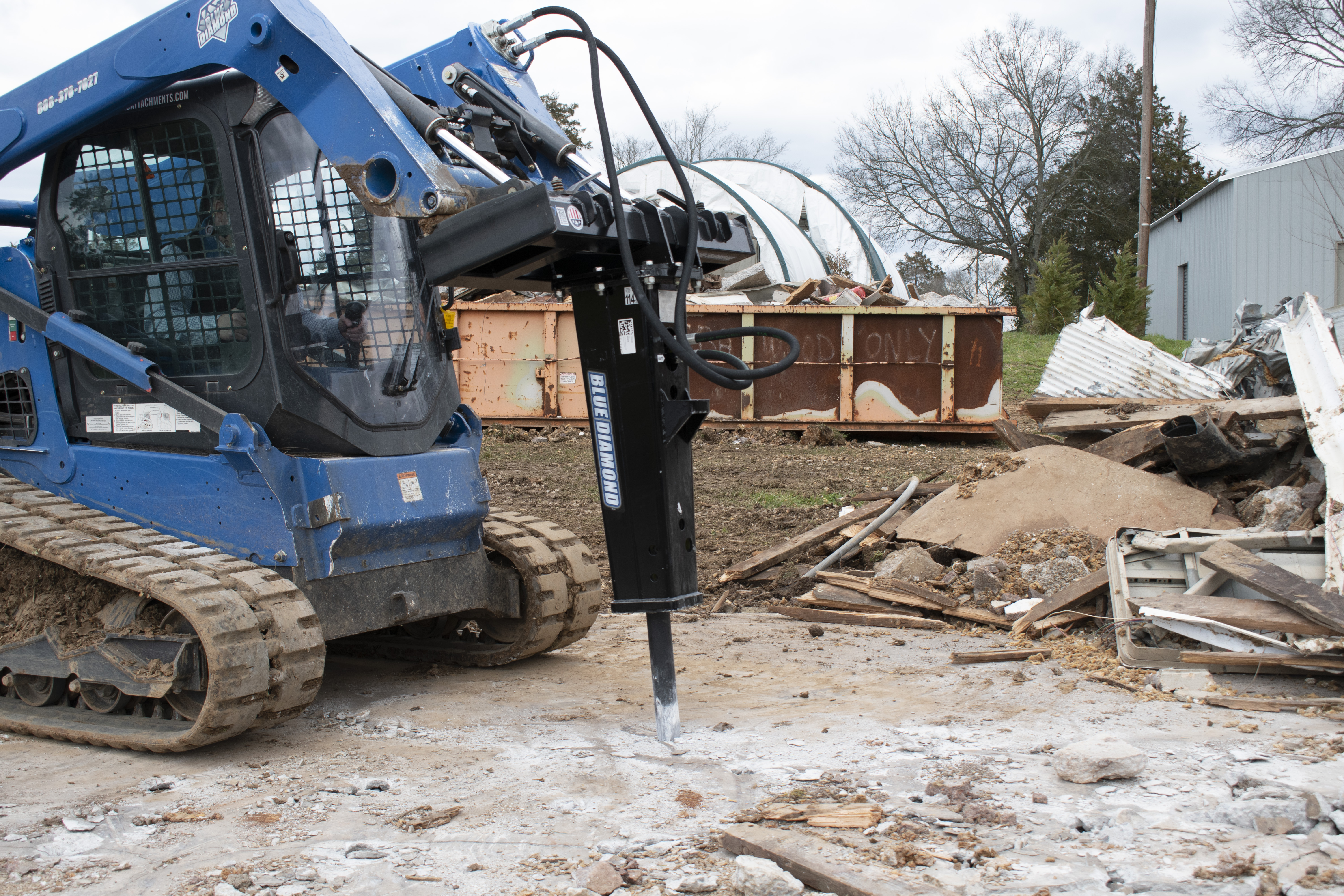 Blue skid steer loader with hydraulic jackhammer attachment at demolition site with debris