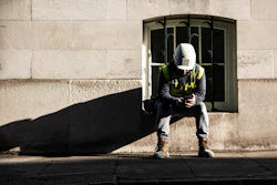 Construction worker in safety gear sitting against stone building wall in sunlight