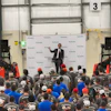 Kubota corporate event with speaker addressing large crowd in industrial warehouse setting with branded machinery