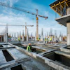 Construction workers in safety vests on concrete foundation with cranes and steel framework at active building site