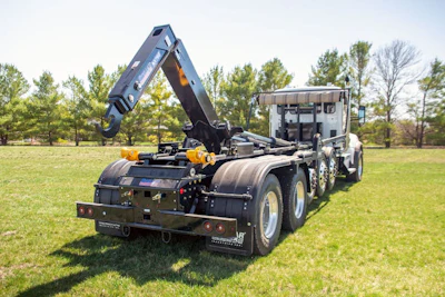 Black and yellow roll-off waste management truck with hydraulic arm system parked in grassy field