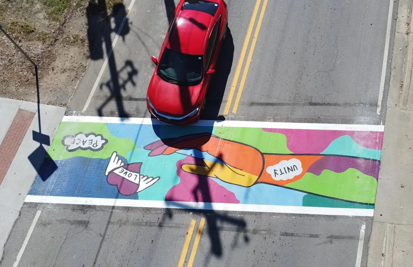Colorful street mural with comic-style graphics and vibrant colors on urban crosswalk, with red car above