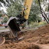 Yellow excavator digging soil at urban construction site surrounded by trees and buildings