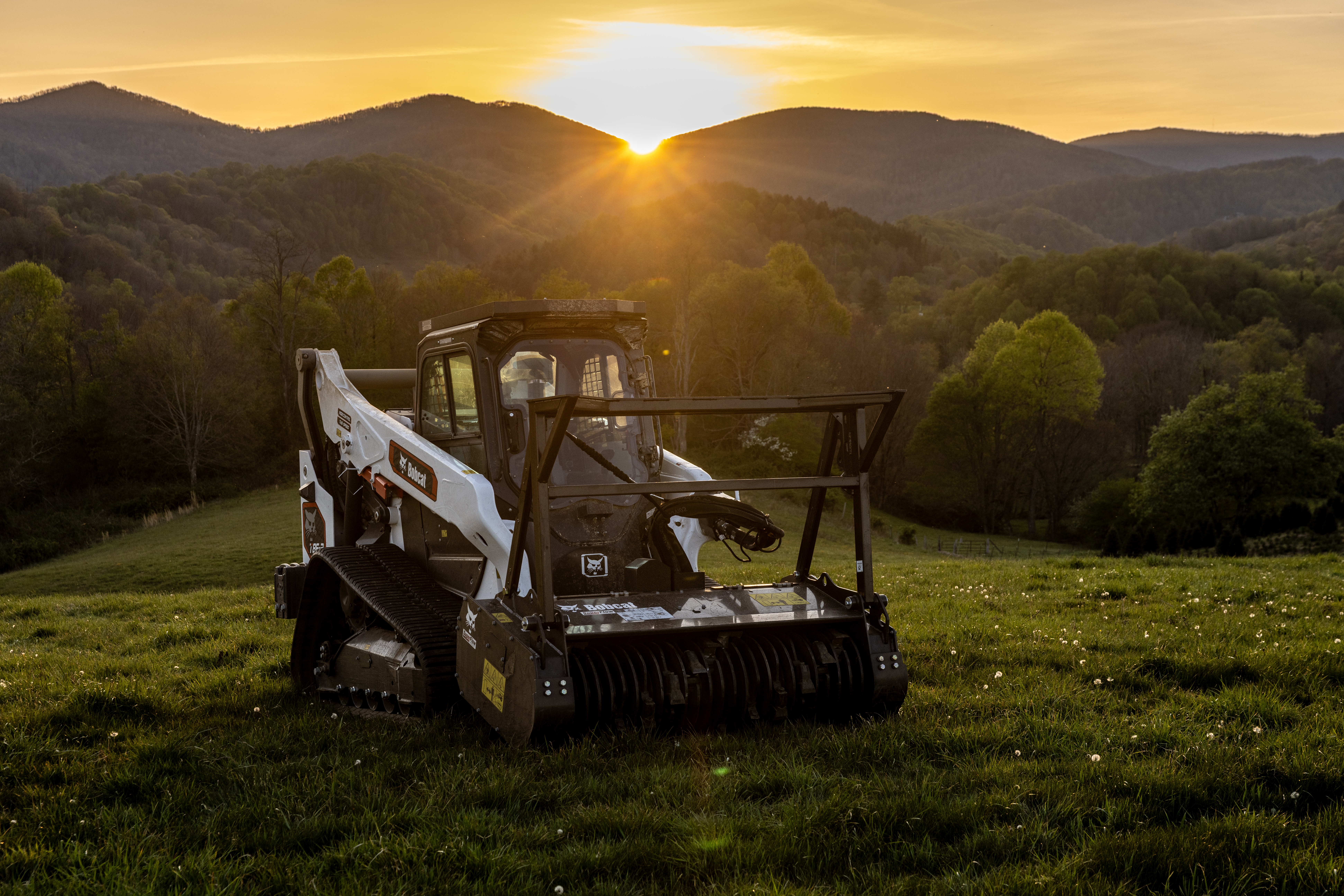 Bobcat skid steer loader positioned in grassy field at sunset with forested mountains in background