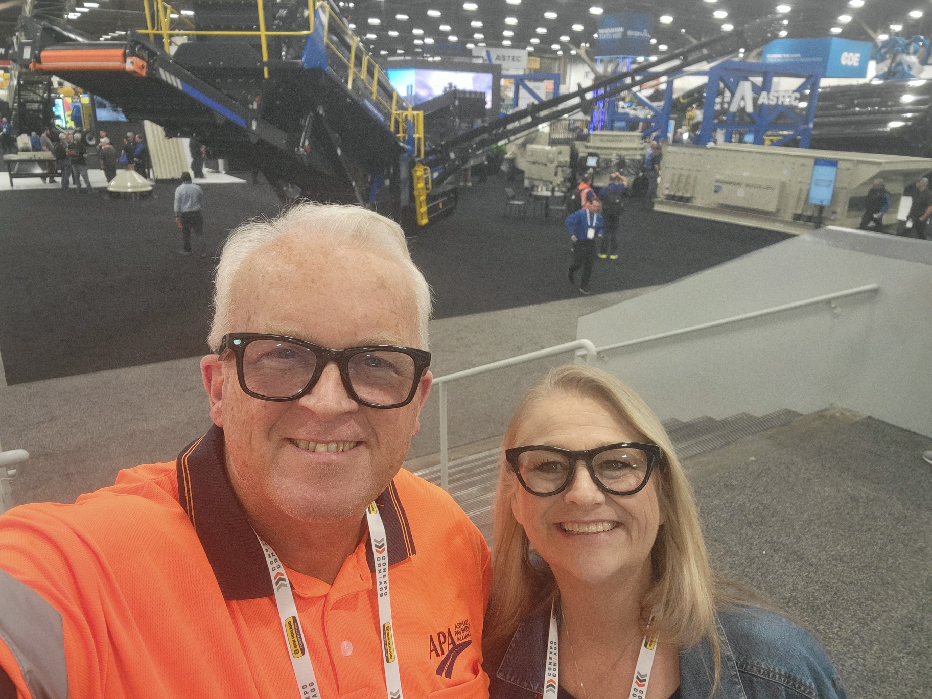 Two people smiling at an industrial trade show with machinery and equipment visible in the background