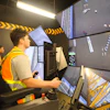Heavy equipment operator training simulator with multiple monitors and yellow safety striping in control room