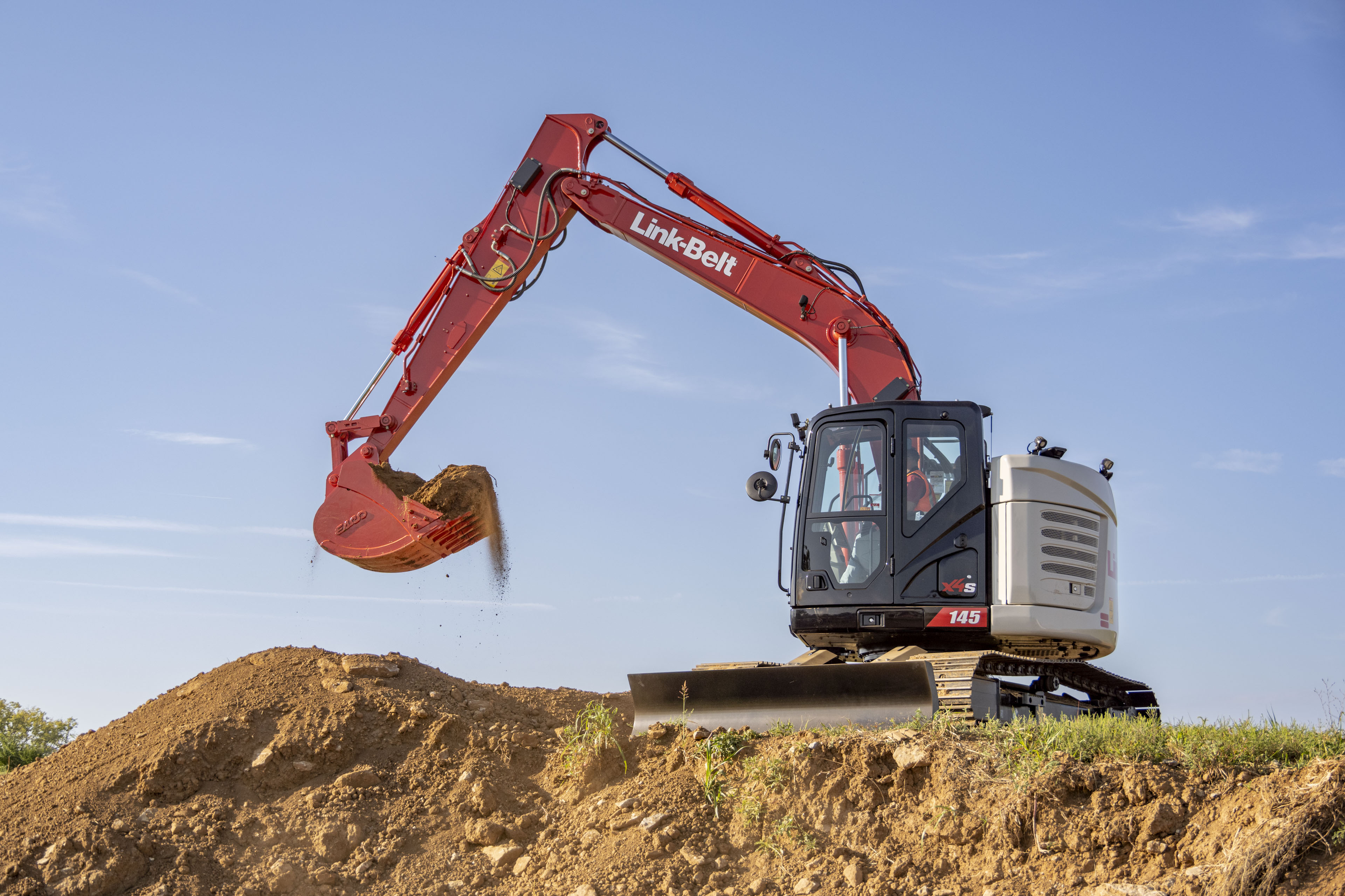 Red Link-Belt excavator with extended boom and bucket digging into dirt pile under blue sky