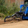 Blue compact track loader with grapple bucket clearing brush and debris from dirt ground near corrugated metal fence