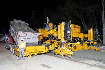 Gomaco concrete paving machine operating at night with workers and dump truck during road construction.
