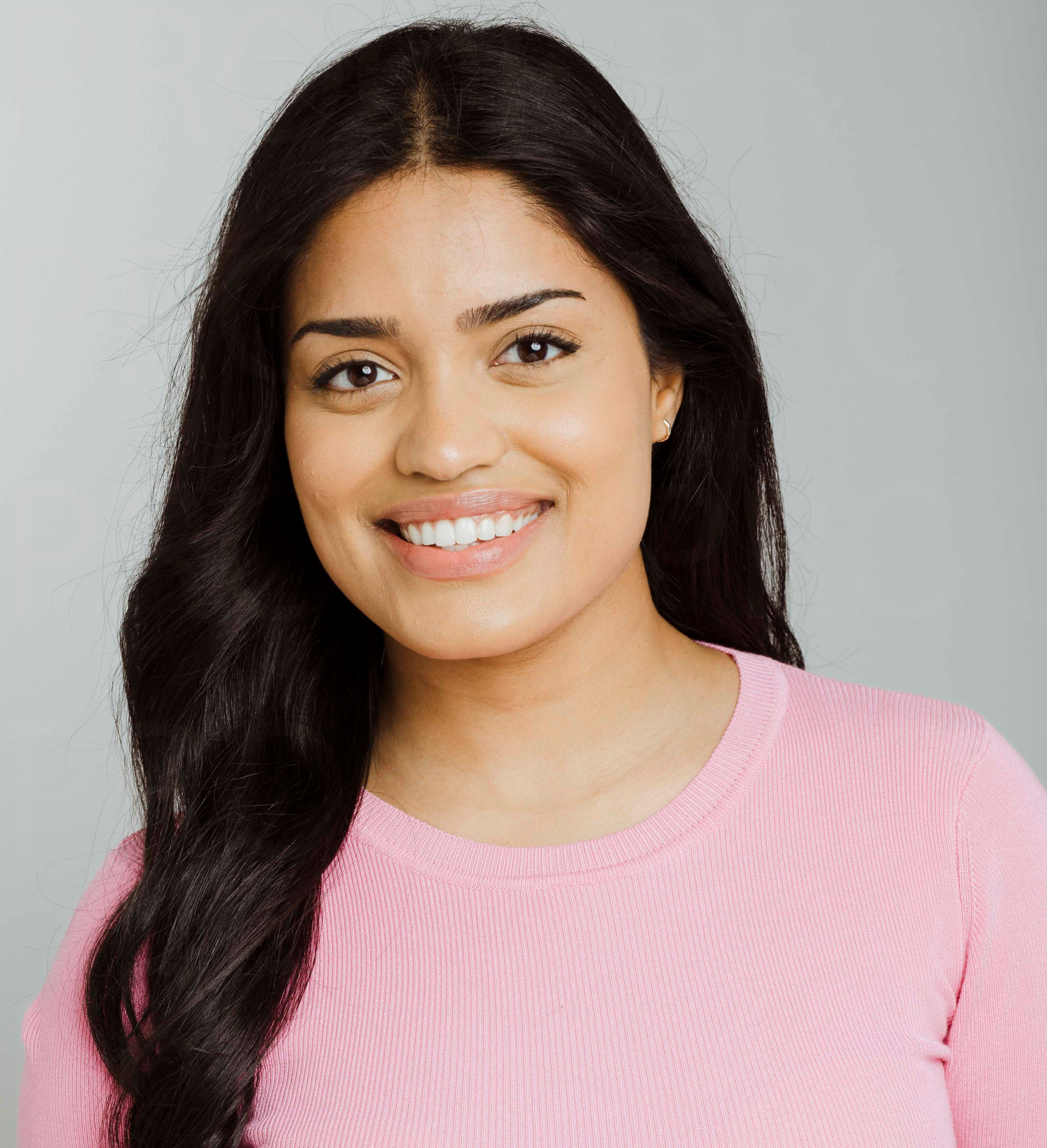 Professional headshot of a smiling woman with long dark hair wearing a pink ribbed sweater against a gray background Headshot
