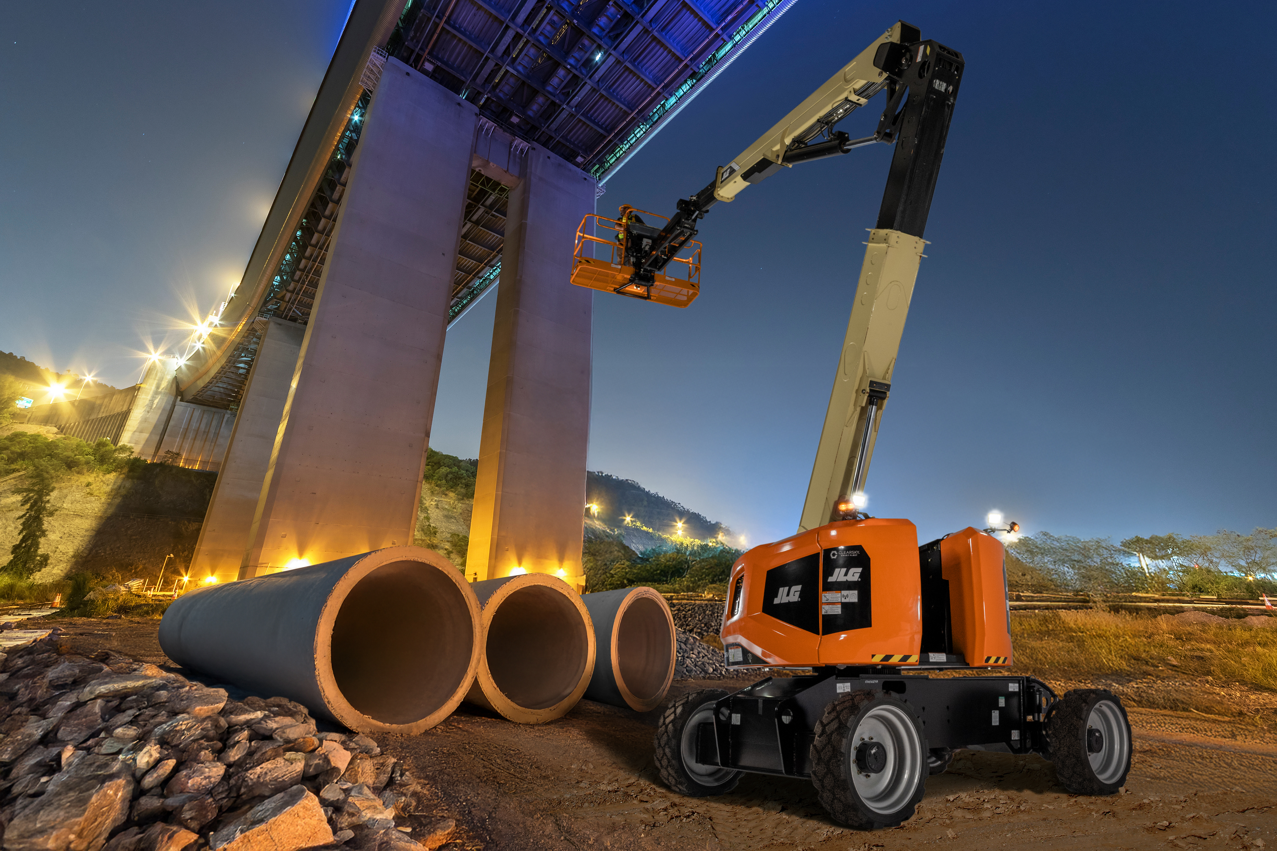 Orange JLG telescopic boom lift operating at dusk near large industrial pipes and concrete structure