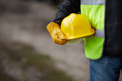 Construction worker holding yellow hard hat wearing neon safety vest and brown gloves