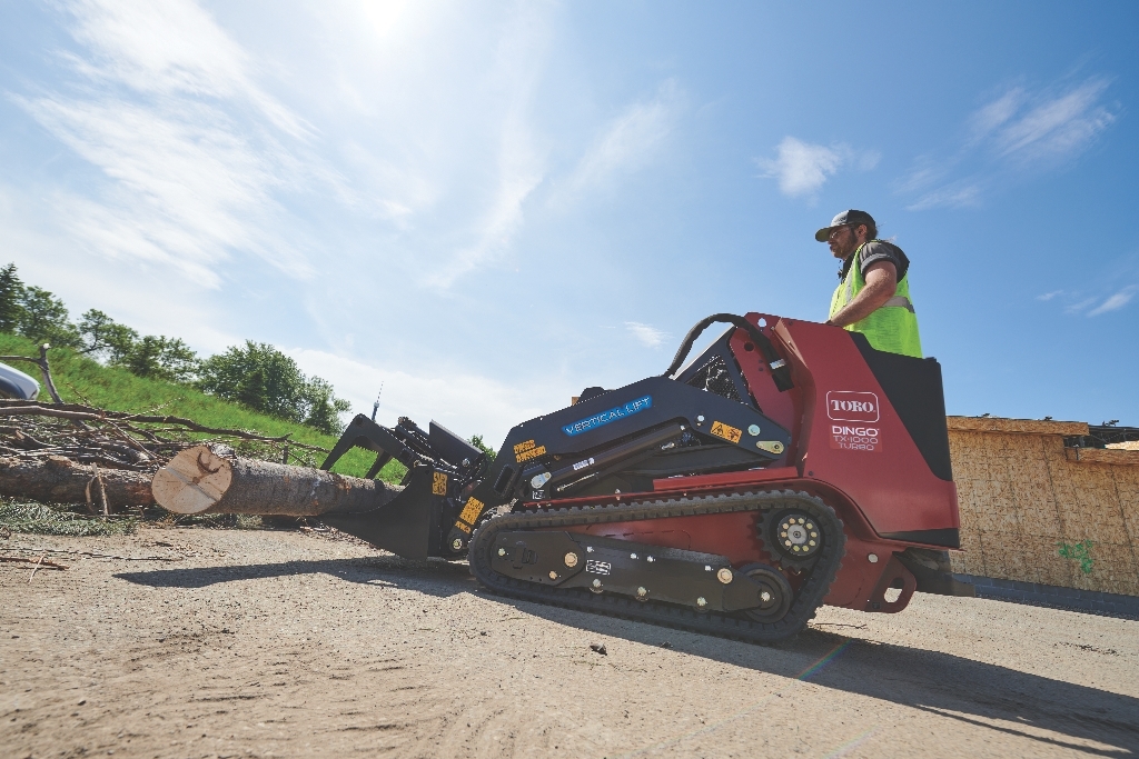 Worker operating a Toro wood chipper on a construction site with logs and materials nearby