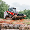 Red and white Bobcat compact track loader with grapple attachment holding large log in wooded area
