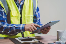 Construction worker in safety vest using tablet at desk with coffee cup and supplies