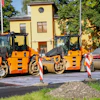 Three orange and black road compactor rollers lined up at a construction site with yellow building in background