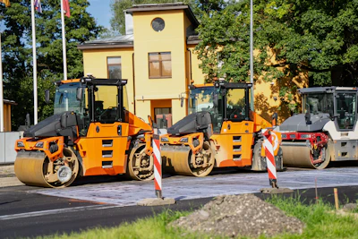 Three orange and black road compactor rollers lined up at a construction site with yellow building in background