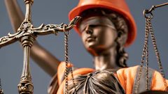 Lady Justice statue wearing orange hard hat and safety vest, holding brass scales against blue sky